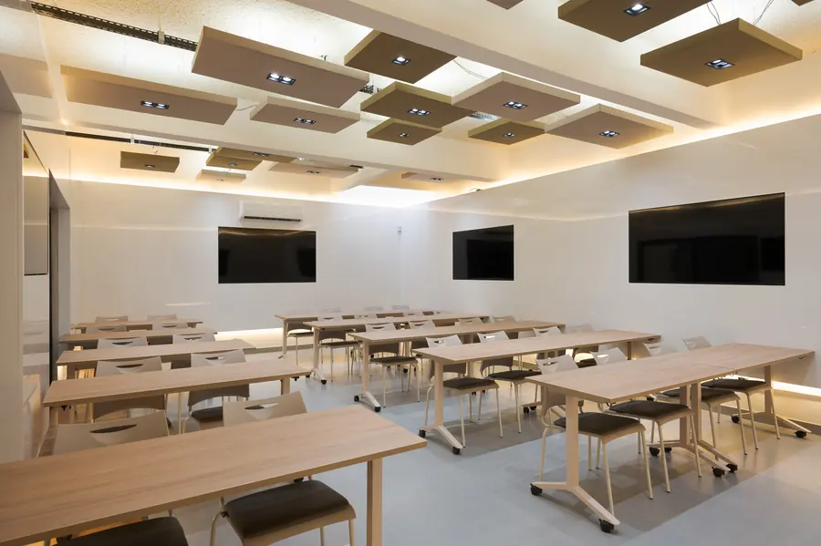 Sala de aula ampla com mesas de madeira, cadeiras e telas nas paredes